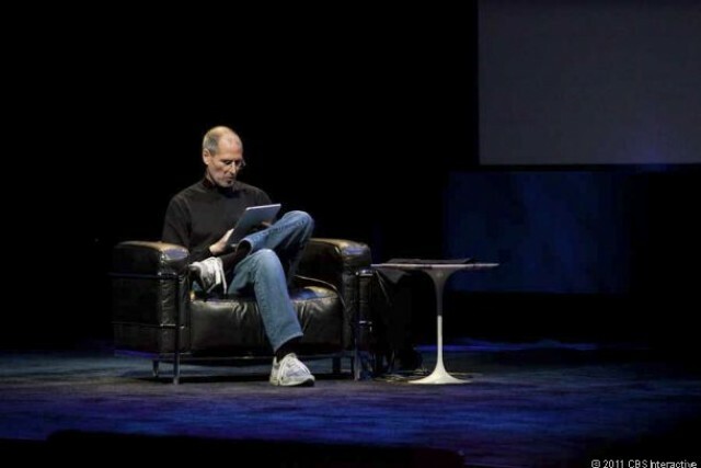 Steve Jobs leunt achterover in een stoel op het podium om te pronken met de iPad.
